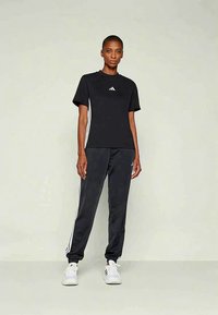 Ženska stoji v črni majici Adidas in hlačah s belimi črtami ter belih supergah Adidas, pred enostavno svetlo ozadje.