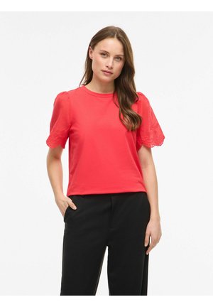Femme portant un chemisier rouge à manches courtes avec des manches en dentelle festonnées et un pantalon noir, debout avec une main dans la poche sur fond blanc.