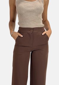 Bruine wide-leg broek met een gladde textuur, hoge taille, voorzakken en een enkele knoopsluiting. Gecombineerd met een geribde beige tanktop.