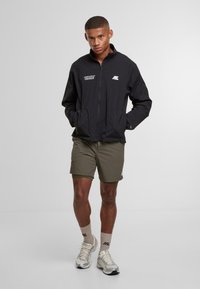 Giovane uomo che indossa una giacca nera con zip, pantaloni corti verde oliva, calze beige e scarpe da corsa argentate, in piedi contro uno sfondo chiaro e semplice.