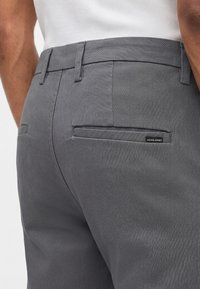 Άνδρας που φορά στενά σκουρόγριμα παντελόνια με πίσω τσέπες και μικρή μαύρη ετικέτα "JACK & JONES" στην δεξιά τσέπη.