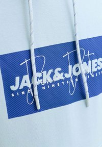 Sweat à capuche blanc avec patch logo rectangulaire bleu et lacets blancs portant le texte "Jack & Jones Since Nineteen" en polices gras et cursive.