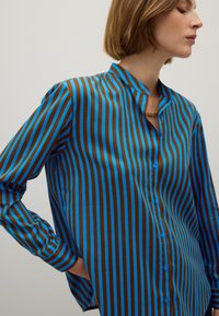 Woman with short hair wearing a blue and brown vertically striped button-up shirt and a gold chain necklace, posing with one hand on her hip.