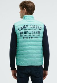 Uomo che indossa un gilet trapuntato azzurro chiaro con la scritta "Camp David Sailing Club Blue Crew" sulla schiena, abbinato a una maglietta a maniche lunghe scura e jeans.