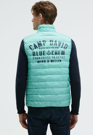 Man wearing light blue quilted vest with "Camp David Sailing Club Blue Crew" text on back, paired with dark long-sleeve shirt and jeans.