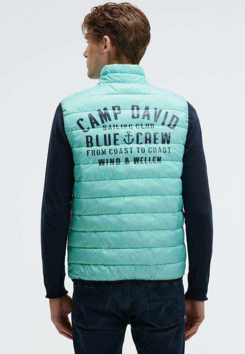 Uomo che indossa un gilet trapuntato azzurro chiaro con la scritta "Camp David Sailing Club Blue Crew" sulla schiena, abbinato a una maglietta a maniche lunghe scura e jeans.