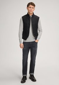 Czarna pikowana kamizelka z wysokim kołnierzem, zapięciem na guziki z przodu i dwoma kieszeniami, noszona na jasnoszarym swetrze i ciemnych dżinsach z podwiniętymi nogawkami. Czarne buty.