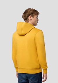 Uomo con i capelli corti castani, che indossa un felpa gialla e jeans blu, di spalle contro uno sfondo grigio chiaro uniforme.