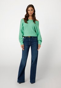 Maglione lavorato a maglia verde menta con design scivolato sulle spalle, polsini a costine, abbinato a jeans a zampa d'elefante blu scuro, creando un outfit rilassato.