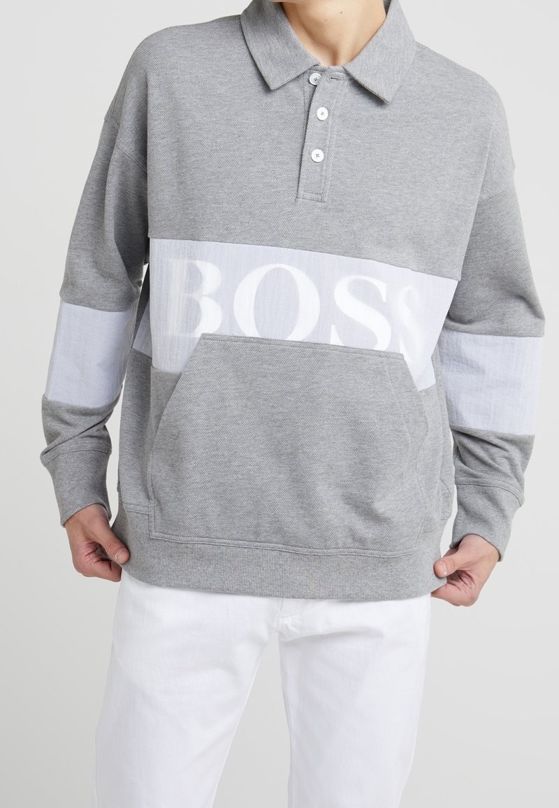 Hombre vistiendo un polo gris de manga larga con una franja horizontal blanca y un gran texto "BOSS", combinado con pantalones blancos.