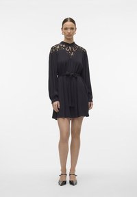 Rochie neagră cu mâneci lungi, detalii din dantelă pe partea superioară a bustului, talie ajustată cu un batic din material și fustă evazată. Stil elegant.