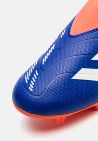 Botín de fútbol con una parte superior texturizada en azul, acentos en blanco y una suela de un naranja brillante. Presenta pequeños puntos en relieve para mayor agarre.