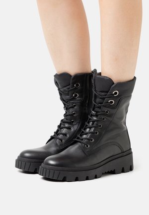 Para czarnych skórzanych, sznurowanych butów bojowych z grubymi podeszwami z bieżnikiem, noszonych na gołych nogach na białym tle.