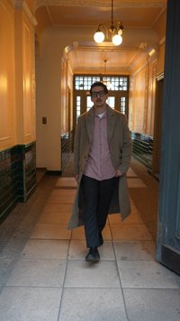 Mężczyzna w okularach, beżowym długim płaszczu, różowej koszuli, czarnych spodniach i butach idzie przez korytarz z płytkami na podłodze, żółtymi ścianami i wiszącym oświetleniem.