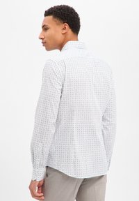 Camicia bianca a maniche lunghe con un piccolo motivo geometrico multicolore, polsini con bottoni e orlo curvo, mostrata dalla parte posteriore.