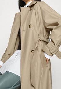 Cappotto trench beige con vita regolata da una cintura, grandi bottoni e spalline. Il tessuto appare morbido, abbinato a una camicia bianca e pantaloni verdi.