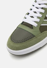 Sneaker verde cu o capacă de vârf din piele netedă și un corp din suede texturat. Șireturi și tălpi albe. Detalii perforate în zona degetelor.