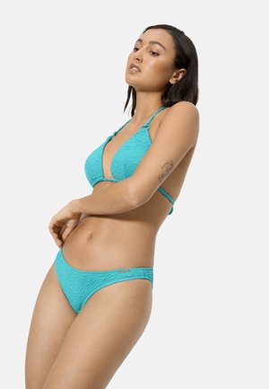 Mujer con tono de piel medio y cabello oscuro que lleva un bikini turquesa texturizado, de pie con una mano cerca de la cintura y mirando de lado.