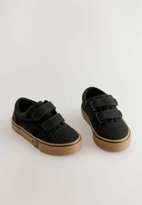 Zwarte canvas schoenen met klittenbandsluitingen, rubberen zolen en een tan buitenzool. Heeft een ronde neus en minimale branding aan de zijkant.