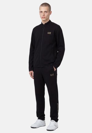 Giovane uomo che indossa una giacca nera EA7 con zip e pantaloni abbinati con sneakers bianche EA7, in piedi con una mano in tasca.