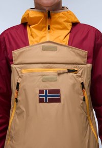 Letvægts vindjakke i beige, rød og gul. Har hætte, lynlåslomme foran og et lille norsk flag patch.