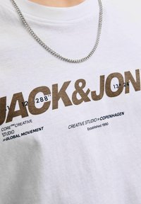 Tricou din bumbac alb cu text grafic maro "JACK & JONES", numere și detalii suplimentare de branding. Lanț argintiu vizibil în jurul gâtului.