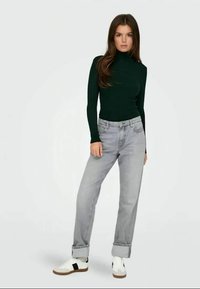 Πράσινη ριγέ ζακέτα με τσέπες, γκρι ψηλόμεσα τζιν και λευκά αθλητικά παπούτσια με καφέ σόλες, σε φόντο απλής υφής.