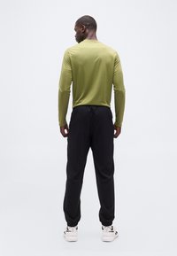 Άνδρας με μακρυμάνικο πουκάμισο ελιάς και μαύρο παντελόνι τζόγκινγκ στέκεται με την πλάτη, φοράει λευκά αθλητικά παπούτσια μπροστά σε απλό λευκό φόντο.