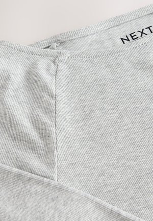 Tessuto grigio chiaro a coste con una texture liscia. Presenta un dettaglio di cucitura e un'etichetta con la scritta "NEXT" stampata in nero. Vista ravvicinata.