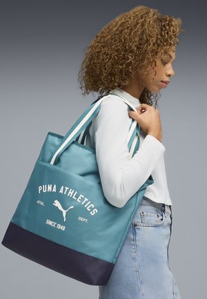 Młoda kobieta o kręconych włosach, ubrana w białą koszulę z długim rękawem i jasne dżinsy, niosąca na ramieniu niebieską torbę typu tote Puma Athletics.