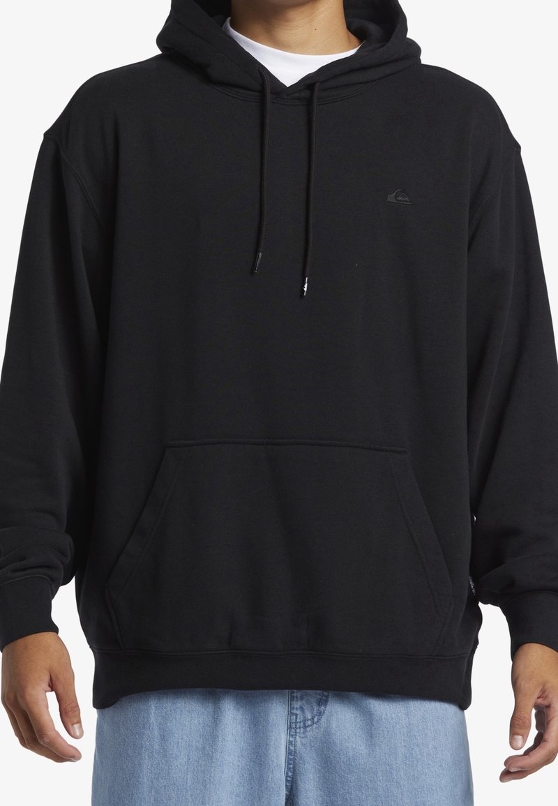 Personne portant un sweat à capuche noir avec poche avant et cordons, associé à un jean bleu clair.