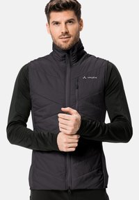 Gilet isolato nero con design a rombi, colletto alto e zip frontale. Indossato sopra una maglia a maniche lunghe, con un logo sul petto.