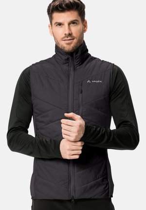 Schwarze isolierte Weste mit einem Karo-gesteppten Design, hohem Kragen und vorderem Reißverschluss. Getragen über einem Langarmshirt, das ein Logo auf der Brust zeigt.