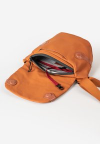 Zaino di stoffa color sabbia con dettagli in pelle. Presenta un compartimento principale con zip e una tracolla rossa regolabile. Texture liscia, design minimalista.