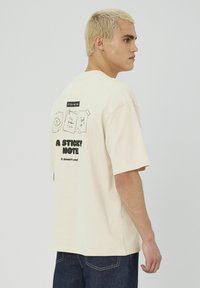 T-shirt oversize de couleur crème en coton, avec des motifs graphiques au dos. L'avant est uni, avec des manches courtes et un col rond.