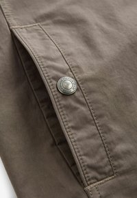 Brunt stof med en glat tekstur, prydet med en sølvknap detalje, der læser "Premium Grade Durable Goods" nær en syet lomme.