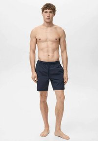 Shorts de bain bleu marine avec taille élastique, longueur mi-cuisse et texture lisse. Modèle debout pieds nus contre un fond neutre.