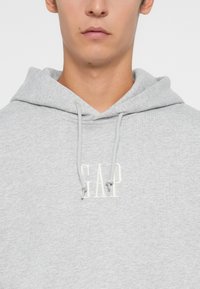 Sudadera de color gris claro hecha de una tela suave, con un bolsillo frontal tipo canguro y un logo blanco de "GAP" en el pecho. Incluye una capucha con cordón.