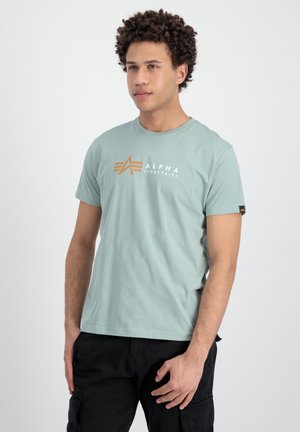 Camiseta de manga corta en verde menta con logo "ALPHA" en blanco y dorado, cuello redondo y etiqueta negra contrastante en la manga izquierda.