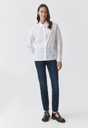 Camicia bianca a maniche lunghe con motivo floreale testurizzato, chiusura con bottoni davanti, abbinata a jeans blu scuro e scarpe nere. Tessuto liscio, vestibilità sartoriale.