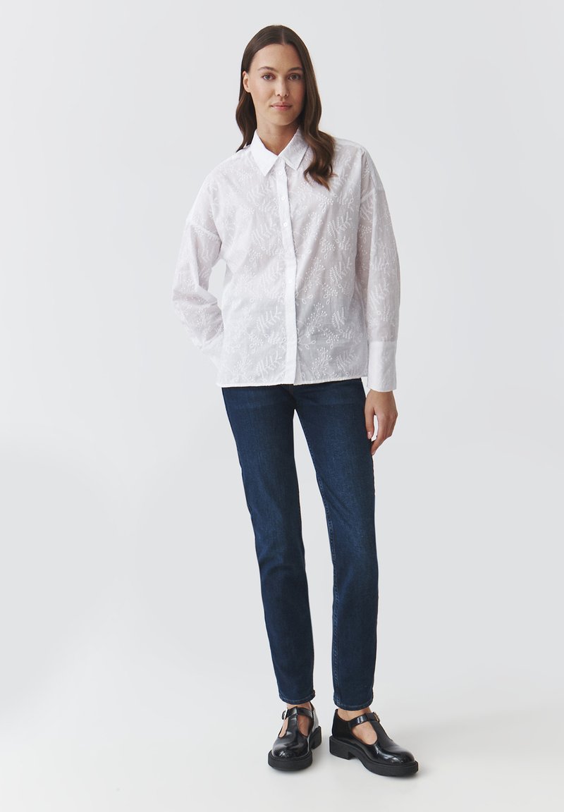 Chemise blanche à manches longues avec motif floral texturé, devant boutonné, associée à un jean bleu foncé et des chaussures noires. Tissu lisse, coupe ajustée.