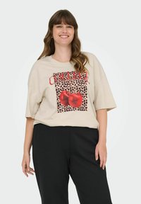 Beżowy, oversize'owy t-shirt z grafiką, z czerwonym napisem "COLLEGE", na tle w panterkę i wzorem z dwoma czerwonymi jabłkami. Czarne spodnie.