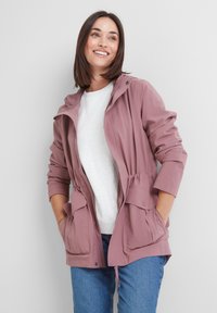 Smiling woman wearing a pink hooded jacket over a white shirt and blue jeans, standing with hands in jacket pockets against a plain background.