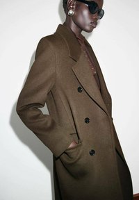 Abrigo de lana marrón de doble botonadura con botones negros y bolsillos laterales. Llevado sobre una camisa oscura, con una textura suave y un diseño a medida.