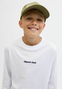 Tricou alb cu mânecă lungă, cu broderie "Original Studio" în negru. Sapcă verde deschis cu logo, având o vizieră curbată. Fața zâmbitoare a unui copil.