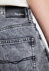 Pilki džinsai su odiniu ženkleliu "Pepe Jeans London" ant juosmens, matoma siuvimo ir kišenių detalė.