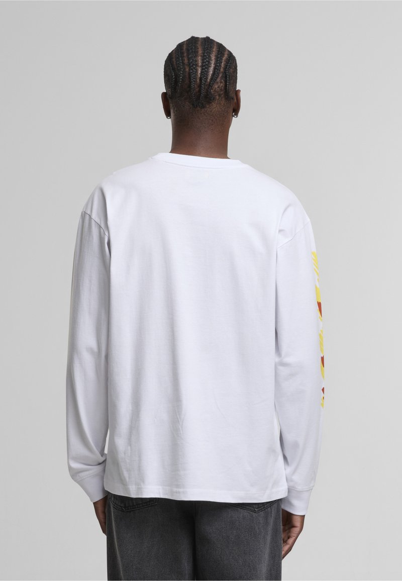 Person with braided hair wearing a plain white long-sleeve shirt and dark pants, shown from the back against a gray background.