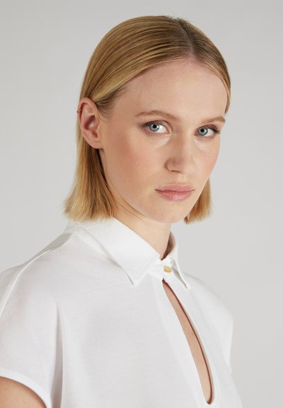 Blusa bianca con colletto, caratterizzata da un dettaglio con bottoni, un'apertura a occhio di bue e maniche corte. Tessuto liscio e leggero.