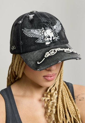 Kobieta z blond warkoczami nosi czarną, zniszczoną czapkę z grafiką czaszki i skrzydeł oraz białym, haftowanym napisem „Ed Hardy” na daszku.