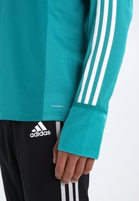 Camicia azzurra a maniche lunghe con design a tre strisce bianche, caratterizzata da un orlo arrotondato e polsini aderenti, abbinata a pantaloni neri con logo Adidas.